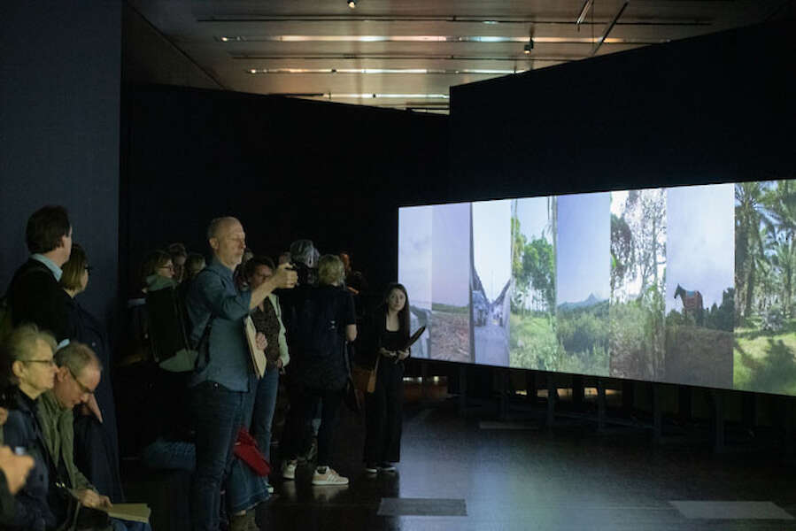 Ausstellungsansicht. Besucher*innen stehen vor einer großen Wand, auf die mehrere hochformatige Videos nebeneinander projiziert werden.