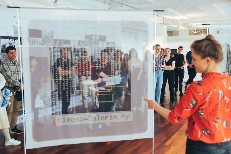 Lilly Lulay steht vor Besuchenden in ihrer Ausstellung. Sie zeigt von hinten auf einen bedruckten Stoff.