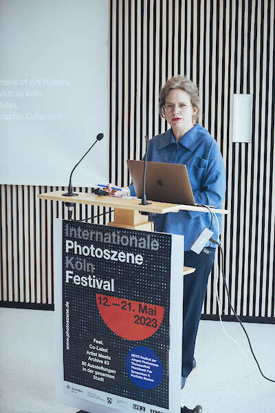 Eine Frau steht an einem Rednerpult. Vor ihr hängt das Poster des Photoszene Festivals 2023.