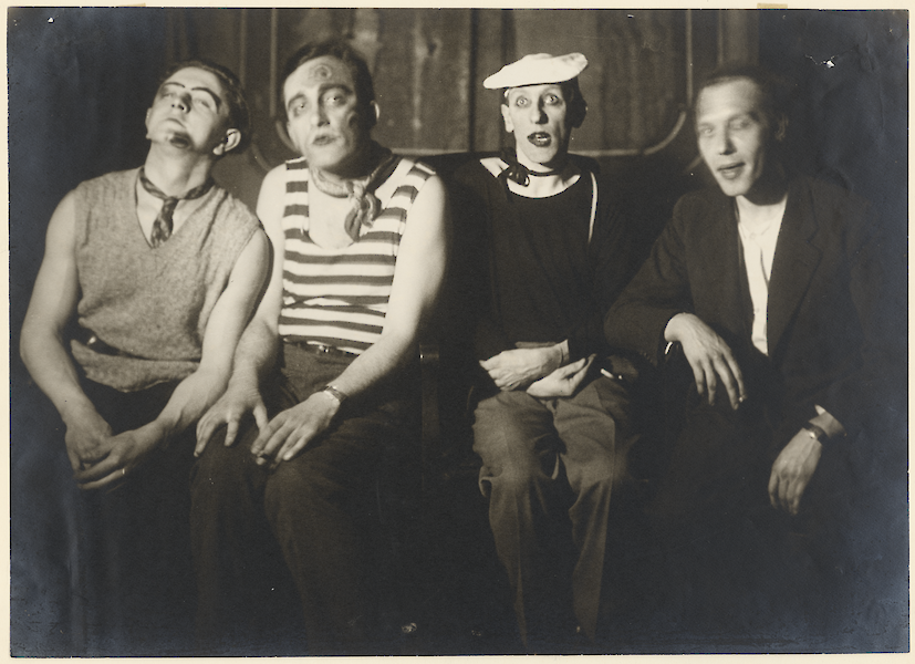 Black and white photo by August Sander. Four artists are sitting in a row, partly made up and disguised.